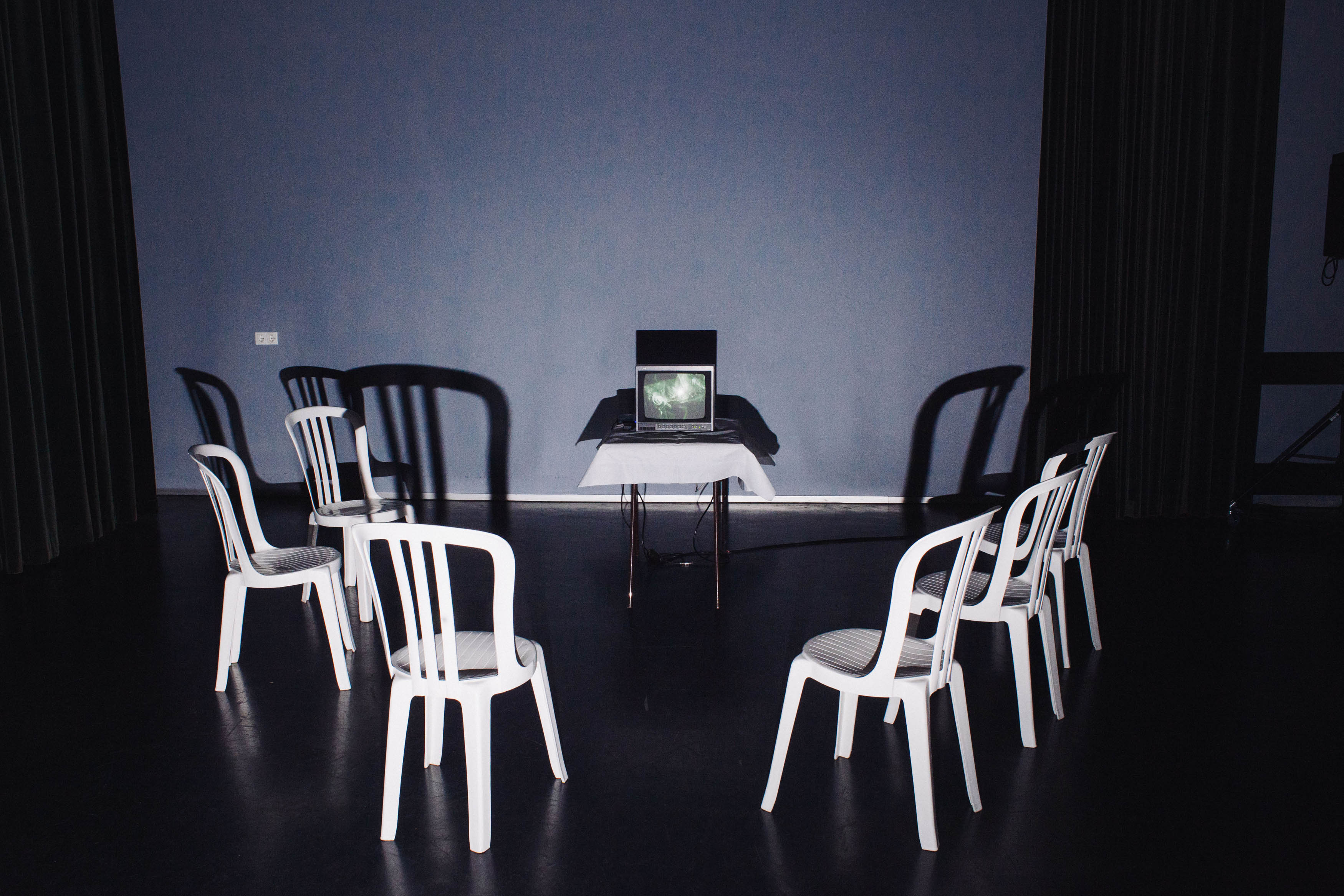 Four white plastic chairs and a small old TV on a table in a rehearsal room. The photo is taken with flash.